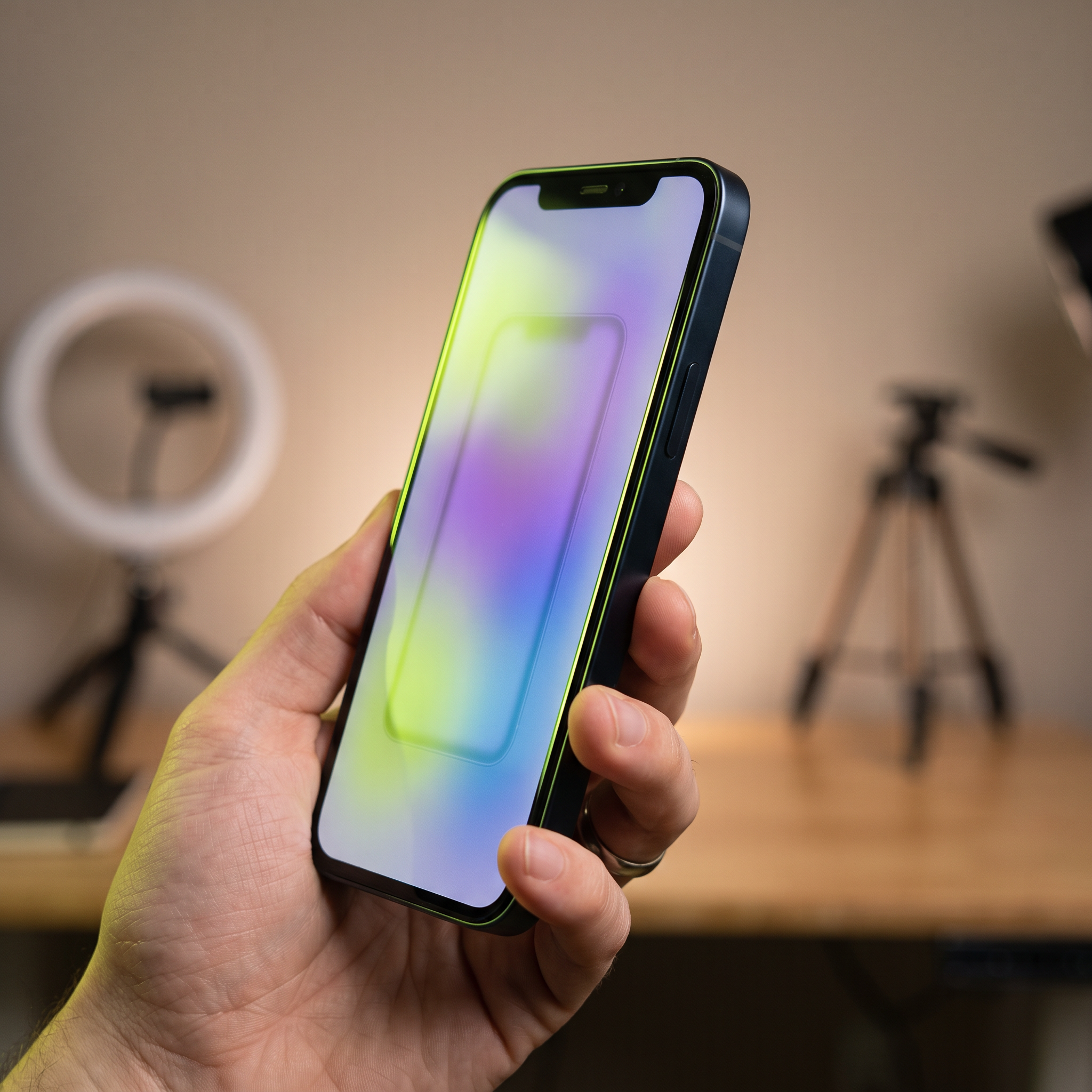A smartphone held vertically in a creator's hand, screen turned at an angle away from direct view so no readable UI appears, showing only a soft blurred gradient glow with a faint silhouette of a vertical video frame; a ring light and phone tripod rig sit softly out of focus behind it